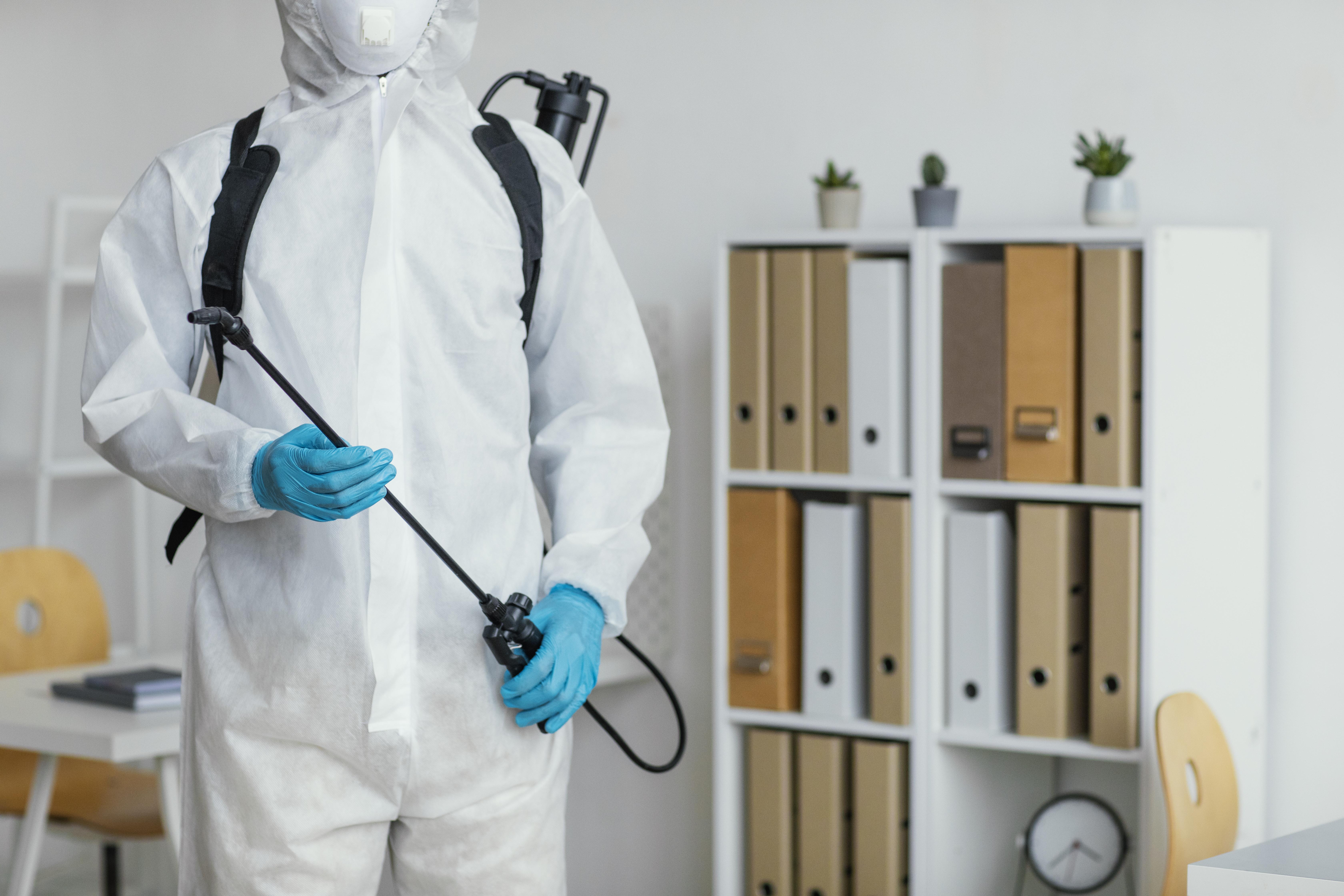 Pest control worker in protective gear with sprayer in an office setting.